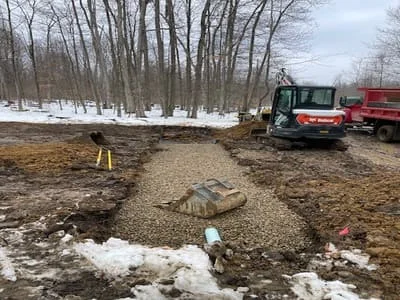 Compact track loader excavating driveway site with gravel base and snow patches visible