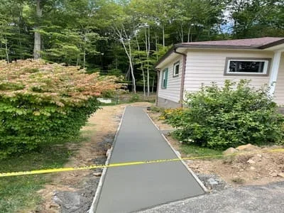Freshly poured concrete walkway with yellow safety line, bordered by landscaping and wooded property