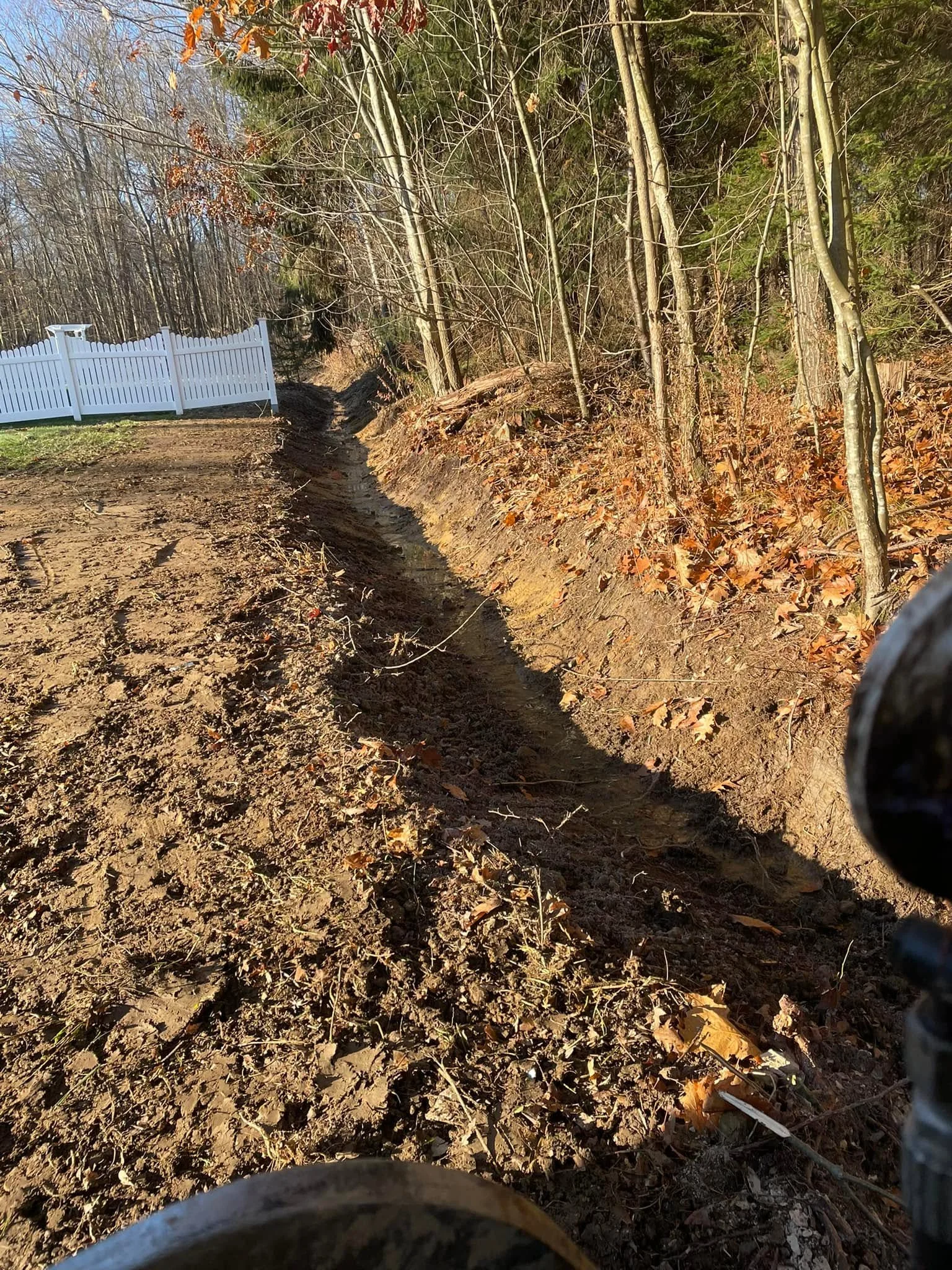 Excavated drainage trench with graded soil banks through wooded area near white vinyl fence